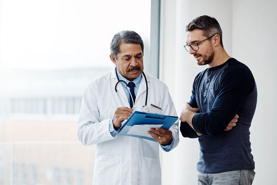 A doctor in a white coat with a stethoscope discusses Hormone Replacement Therapy notes on a clipboard with a man in glasses and casual clothing beside a large window in a bright room.