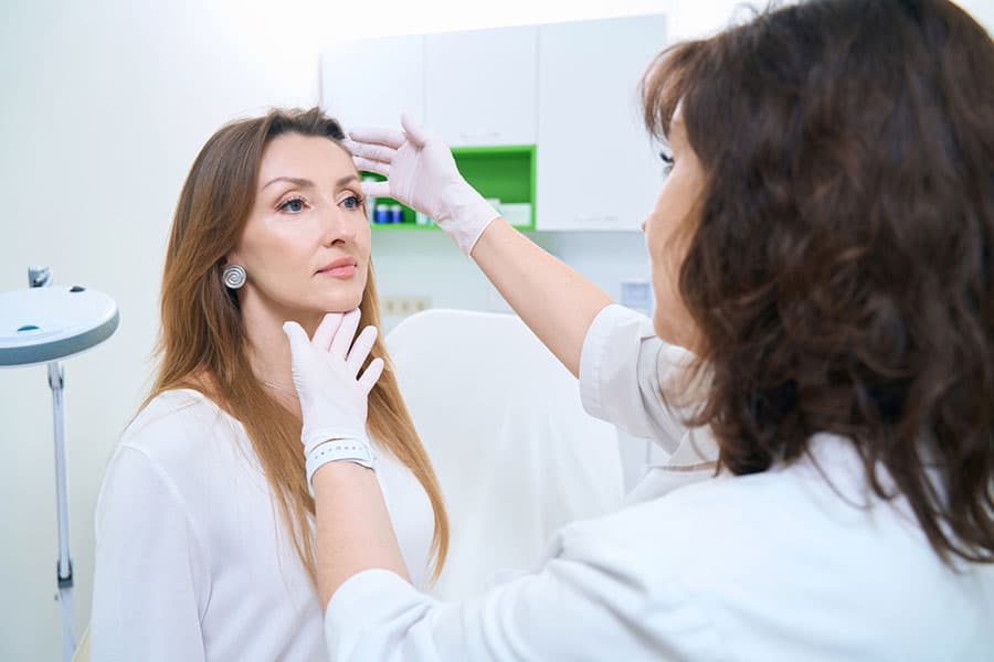 A female doctor wearing gloves examines a woman's face in a medical office, possibly assessing her skin or preparing for a science-backed cosmetic procedure. The patient sits calmly and looks ahead.