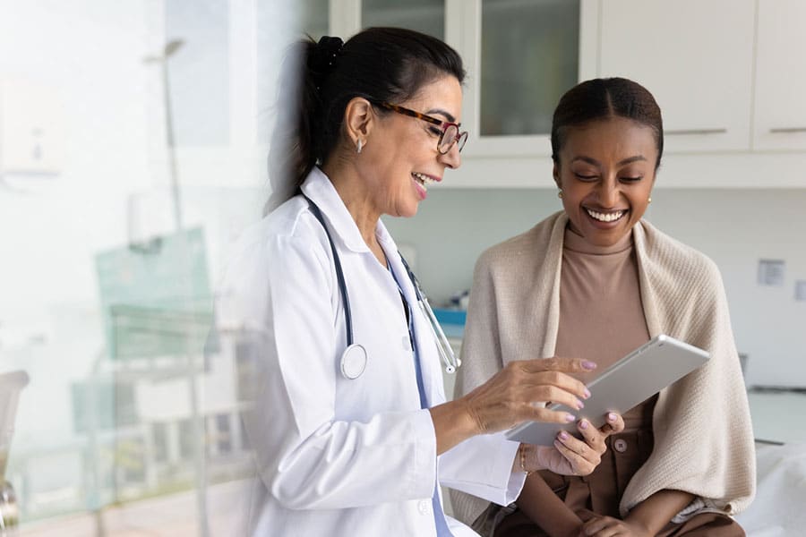 A doctor in a white coat shows a tablet to a smiling patient sitting beside her in a Dearborn medical office, discussing PRP Treatment. Both appear engaged and happy during their conversation.