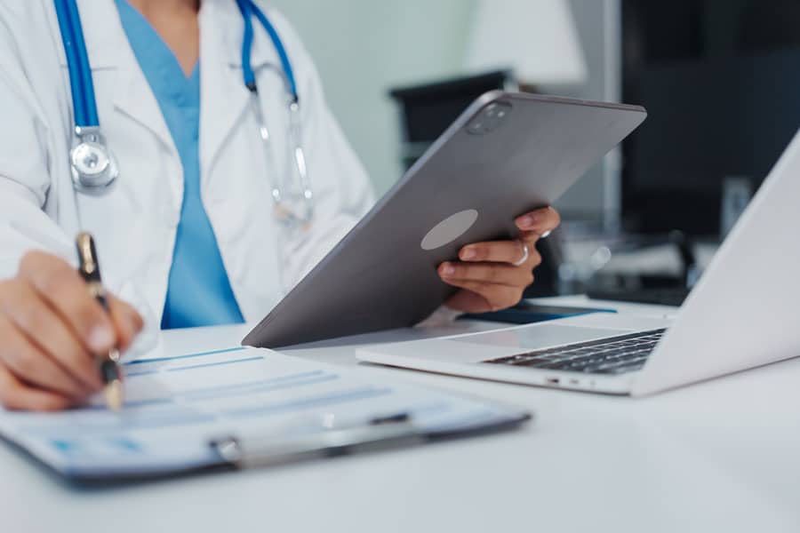 A doctor in a white coat sits at a desk in St. Clair Shores, holding a tablet and writing on a clipboard, reflecting advancements in Regenerative Medicine alongside his laptop.