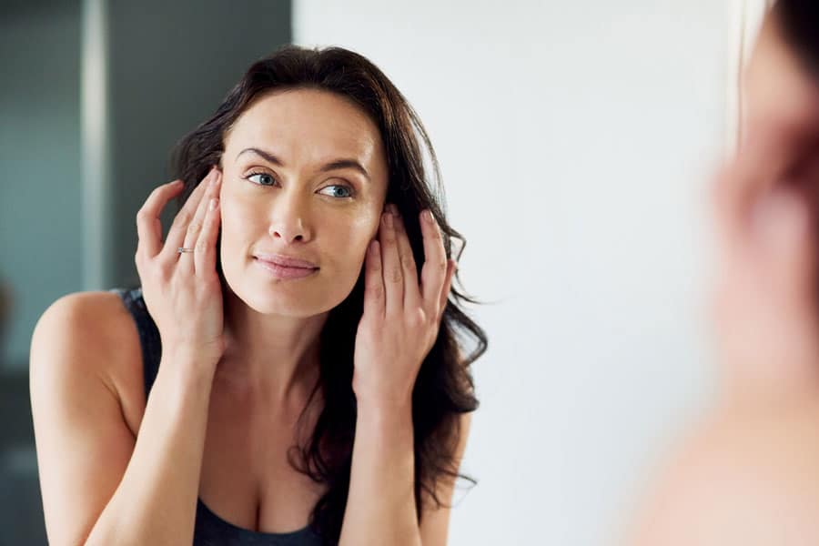 A woman with long dark hair looks at herself in the mirror, gently touching her face with both hands, wearing a gray tank top in a softly lit room—reflecting on the reduced inflammation and faster healing from PRP therapy.
