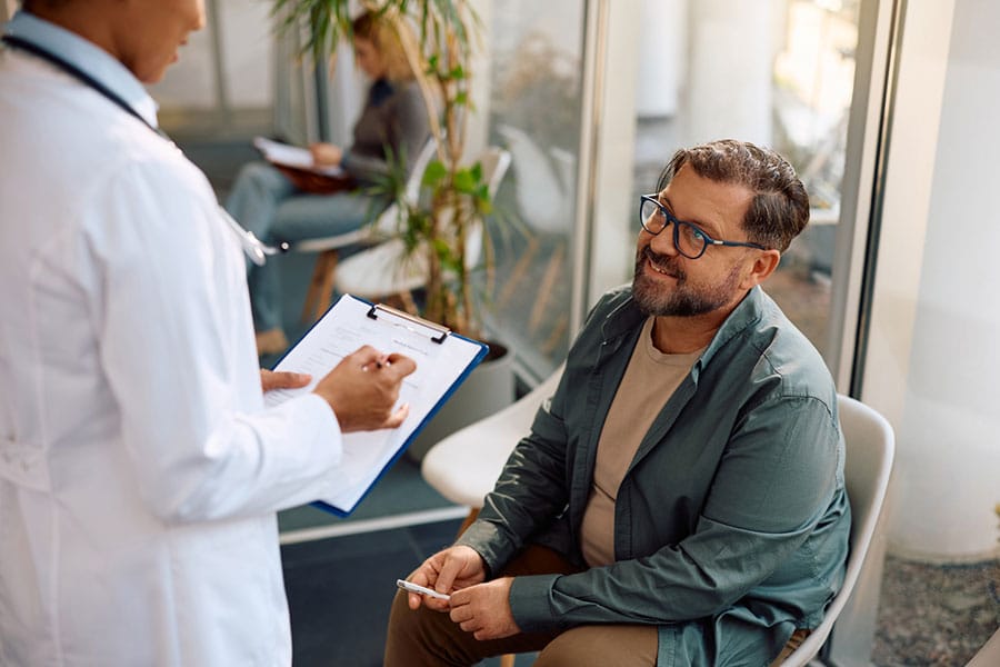 A man with glasses sits in a waiting area, smiling up at a doctor holding a clipboard. Another person sits in the background. The setting appears to be a modern medical office with large windows and plants, suggesting an environment where innovative treatments like regenerative medicine might be discussed or explained—prompting patients to ask, “What is regenerative medicine?”.