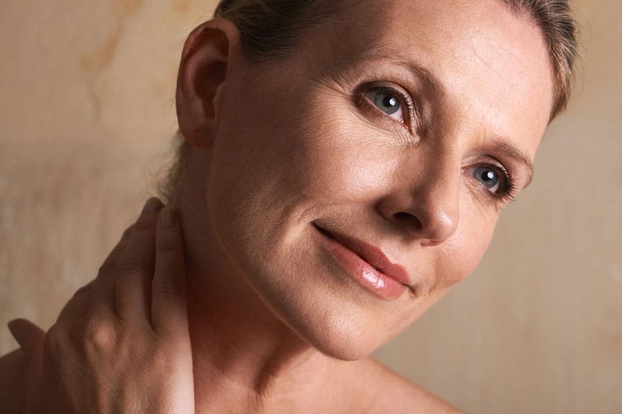 A middle-aged woman with light skin and blue eyes smiles softly, looking slightly upward. She gently touches her neck with one hand, embracing beauty and health, as the background is softly lit in warm tones.