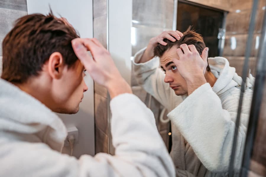 A person in a white bathrobe stands in front of a mirror, using both hands to inspect their scalp and hair for signs of hair loss while looking closely at their reflection in a bathroom.