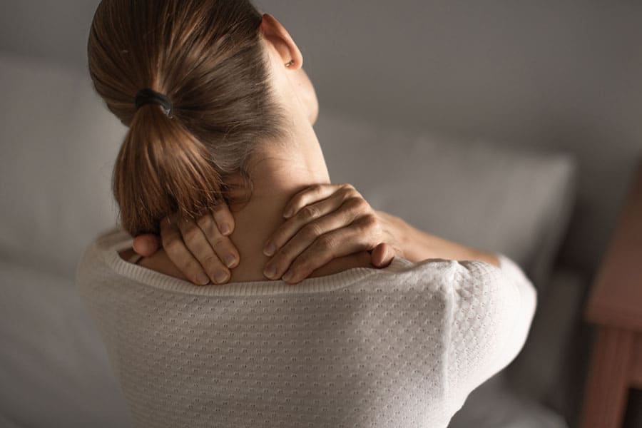A woman with brown hair in a ponytail sits on a bed, holding the back of her neck with both hands, as if experiencing neck pain or discomfort—possibly making her a candidate for regenerative medicine treatments.