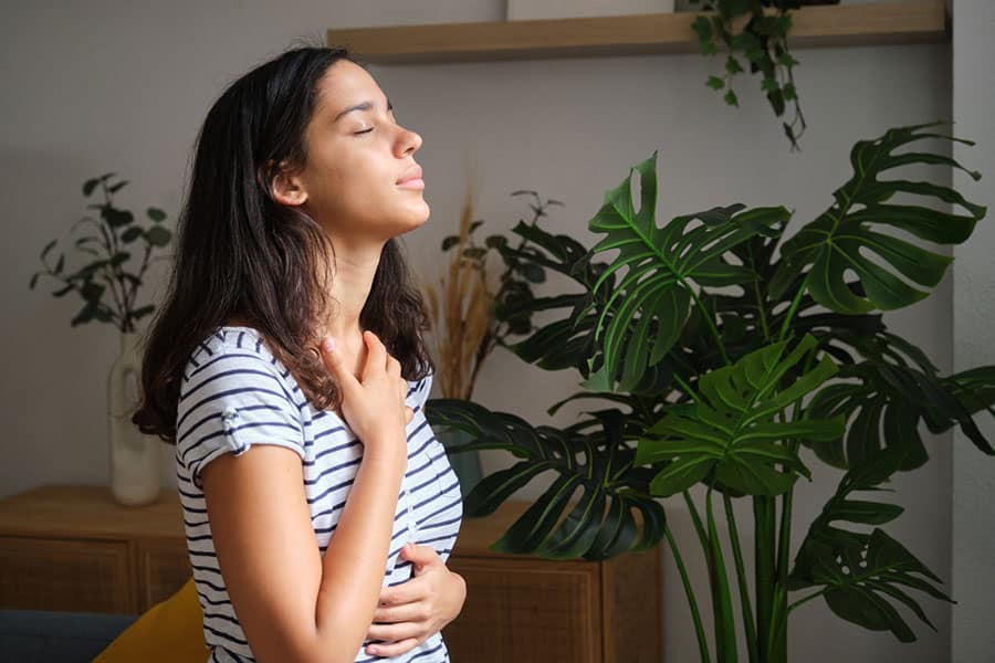 A woman in a striped shirt sits with her eyes closed, breathing deeply beside a large monstera plant while reflecting on her progress with testosterone replacement therapy.
