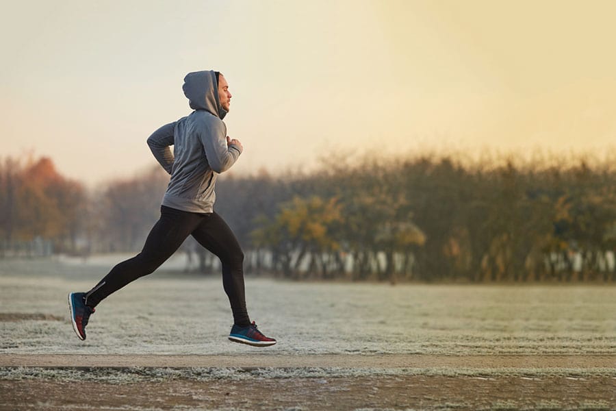 A man in a hooded sweatshirt and leggings jogging on a path at sunrise, reflecting the active lifestyle goals of those on testosterone replacement therapy.